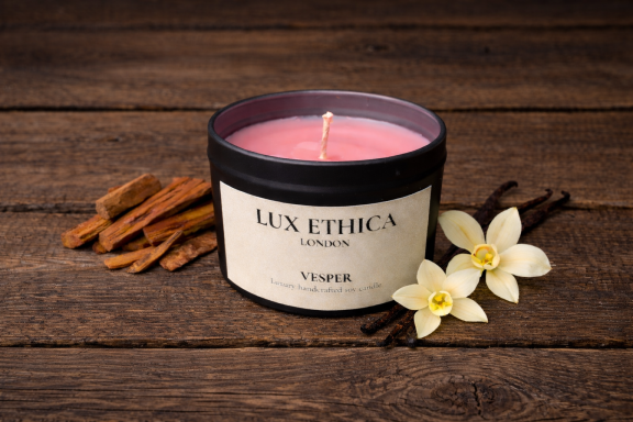 Vesper Pink candle in a black container surrounded by cinnamon sticks and flowers on a wooden surface.
